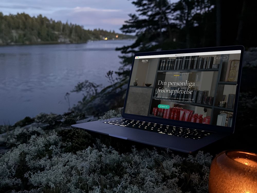 ikonfrisorerna.se öppen i en bärbar dator på en lavklädd klippa vid vattnet i skymningen, med en tänd lykta bredvid.