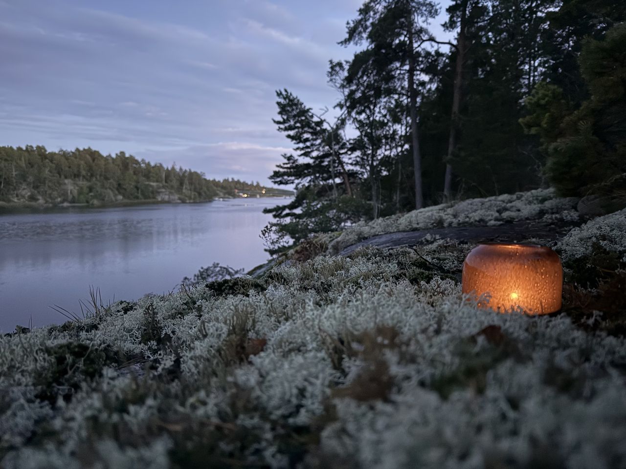 En tänd lykta på en lavklädd klippa vid vattnet i skymningen, med tallar och skogsklädda holmar på andra sidan sundet.