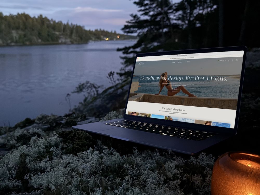 oceiathelabel.com öppen i en bärbar dator på en lavklädd klippa vid vattnet i skymningen, med en tänd lykta bredvid.